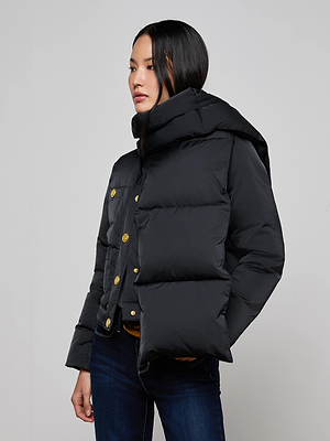 A woman models the Juana Scarf Puffer Jacket—glamorous and black with a large hood—paired with dark jeans and a black button-up shirt, standing against a plain light gray background and gazing slightly to the side.