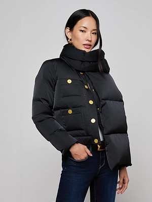 A woman in the Juana Scarf Puffer Jacket with gold buttons and a detachable scarf stands against a plain background. She has straight black hair, gold hoop earrings, dark jeans, and one hand in her pocket.