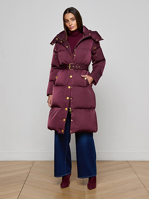 A woman stands against a plain light gray wall, wearing the L'AGENCE Bryson Maxi Puffer Coat with gold buttons and down filling, styled with a maroon turtleneck, wide-leg blue jeans, and matching maroon ankle boots.