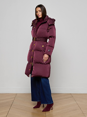 A woman stands indoors on a wooden floor, facing forward with one hand in her pocket, wearing the Bryson Maxi Puffer Coat by L'AGENCE over blue wide-leg jeans and maroon heeled boots against a plain background.