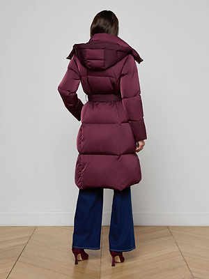 A woman stands facing away in the Bryson Maxi Puffer Coat by L'AGENCE, a burgundy down-filled, belted coat. She pairs it with wide-leg blue jeans and heeled burgundy shoes on a wooden floor against a plain light wall.