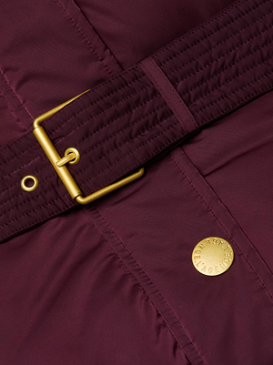 Close-up of maroon quilted fabric from the Bryson Maxi Puffer Coat by L'AGENCE, with down filling, a matching belt with gold buckle, and a round gold button embossed with letters.