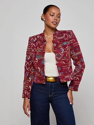 A woman wears the L'AGENCE Asha Jacquard Jacket in red with ruffled edges, paired with a white tank top, dark blue high-waisted jeans, and a black belt featuring a large gold buckle, standing against a plain light gray background.