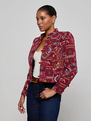 A woman with slicked-back hair wears the L'AGENCE Asha Jacquard Jacket in red over a white top and dark jeans, standing before a plain light background and facing left with a confident expression.