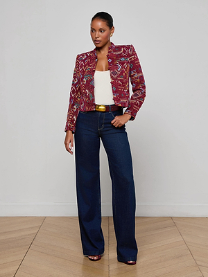 A woman poses confidently on a wooden floor against a light wall, wearing the L'AGENCE Asha Jacquard Jacket over a white top, styled with dark wide-leg jeans, a gold-buckled belt, and heeled sandals.