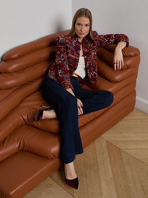 A woman wearing the L'AGENCE Asha Jacquard Jacket sits on a modern brown sofa, one leg crossed, paired with a white top, dark jeans, and burgundy shoes. Her hand rests on the armrest as she looks directly at the camera.