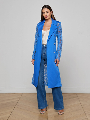 A woman with long brown hair poses indoors against a plain light background, wearing the Amata Lace Trench Coat in regatta blue over a white satin top and high-waisted blue jeans, styled with silver heels on a wooden floor.