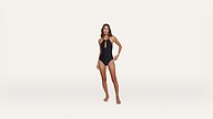 A woman stands barefoot against a plain white background, wearing a black, one-piece swimsuit with a keyhole cutout at the chest and thin straps.