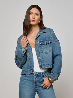 A woman with straight brown hair wears the L'AGENCE Vianney Jean Jacket over a white top and blue jeans. She accessorizes with gold earrings and a bracelet, posing with one hand on her collar against a neutral background.