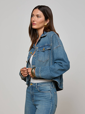 A woman with long brown hair wears the L'AGENCE Vianney Jean Jacket and matching relaxed-fit jeans, standing against a plain light background. She accessorizes with large gold hoop earrings and gold bangles, looking to the side.
