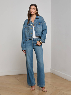 Indoors, a woman wears the L'AGENCE Vianney Jean Jacket over a white top with relaxed blue jeans, gold hoop earrings, a chunky bracelet, and beige open-toe heels on a wooden floor.
