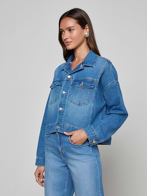 A woman with long brown hair models the Vianney Jean Jacket and matching blue jeans, standing against a plain light gray background with her hands in her jacket pockets, gazing slightly to the side.