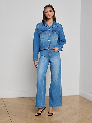 A woman with long brown hair poses indoors against a light wall and wooden floor, wearing the Vianney Jean Jacket in a light wash with matching wide-leg jeans and black high-heeled sandals.