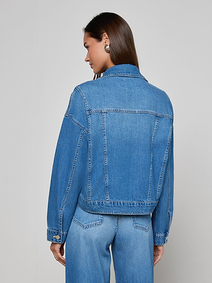 A woman with long brown hair, wearing large round earrings, faces away from the camera in a Vianney Jean Jacket and matching blue jeans against a plain light background.