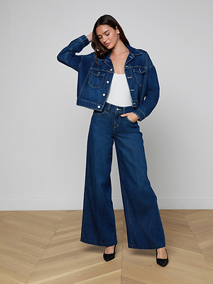A woman with long brown hair stands indoors on a wooden floor, wearing the Vianney Jean Jacket in indigo wash with matching wide-leg jeans, a white top, and black shoes. She faces forward with a neutral expression.