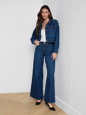 A woman with long brown hair sits on a black leather chair, confidently looking at the camera while wearing the Vianney Jean Jacket in indigo wash paired with wide-leg jeans and black heeled boots.