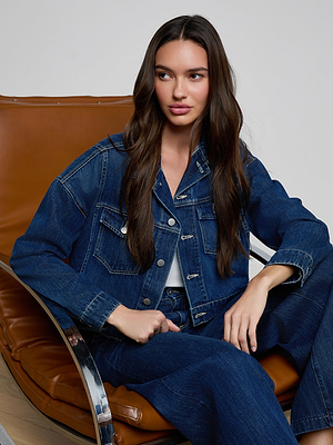 Wearing the Vianney Jean Jacket over a white top, a woman with long brown hair sits on a brown leather chair in matching jeans, looking slightly to the side with a neutral expression.