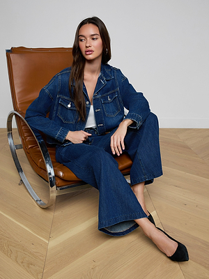 A woman in the Vianney Jean Jacket and wide-leg jeans sits on a modern brown leather and metal chair on a light wood floor. She has long brown hair, wears black heels, and gazes slightly to the side.