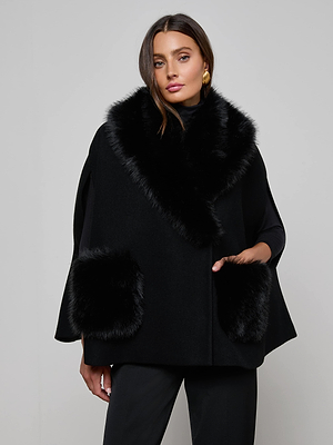 A woman with long, dark hair and gold earrings wears the L'AGENCE Chana Faux Fur Cape—a black cape crafted from a recycled wool blend with faux fur accents—standing confidently against a plain gray background.