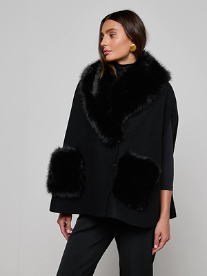 A woman wears the L'AGENCE Chana Faux Fur Cape, featuring faux fur at the collar and pockets and crafted from a recycled wool blend, styled with black pants and gold earrings against a plain light gray background.