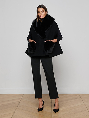 A woman poses against a plain background wearing the L'AGENCE Chana Faux Fur Cape—a modern update crafted from a recycled wool blend—paired with black pants, heels, gold earrings, and straight brown hair.