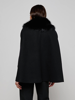 A person with straight brown hair is seen from the back, wearing the L'AGENCE Chana Faux Fur Cape and black pants against a plain light background. The cape features a wide faux fur collar and is made from a recycled wool blend.