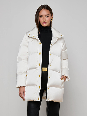 A woman stands against a plain background wearing the L'AGENCE Loretta Puffer Coat in ivory with gold buttons, paired with a black turtleneck and black pants. She has one hand in her pocket and looks at the camera.