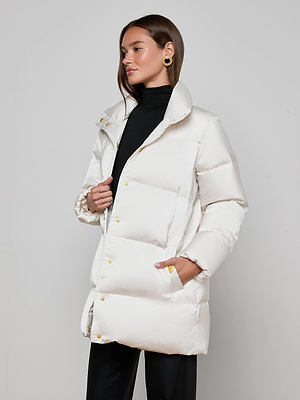 A woman in the L'AGENCE Loretta Puffer Coat, an ivory jacket with gold buttons, wears it over a black turtleneck and pants while standing against a gray background, looking to the side with one hand in her pocket.