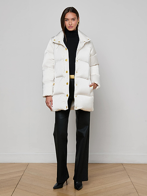 A woman stands against a plain wall wearing the L'AGENCE Loretta Puffer Coat in ivory with gold buttons, paired with a black turtleneck, wide-leg pants, and pointed-toe heels. She faces the camera with a neutral expression.