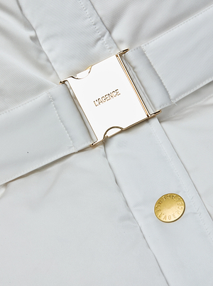Close-up of the Loretta Puffer Coat by L’AGENCE: an ivory quilted jacket with a shiny gold buckle clasp engraved with L’AGENCE, a matching gold button, and a standout statement belt. The smooth fabric completes this elegant look.