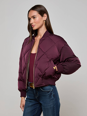 A woman in the L'AGENCE Violeta Reversible Puffer, a maroon satin bomber jacket, with a matching top and blue jeans stands against a light gray background, posing with one hand on her hip. She has long brown hair.