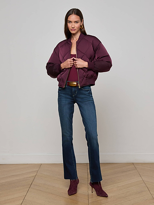 Indoors, a woman poses on a light wood chevron floor wearing the L'AGENCE Violeta Reversible Puffer in maroon satin, paired with dark blue jeans and matching maroon heeled boots, hands holding the front of her jacket.