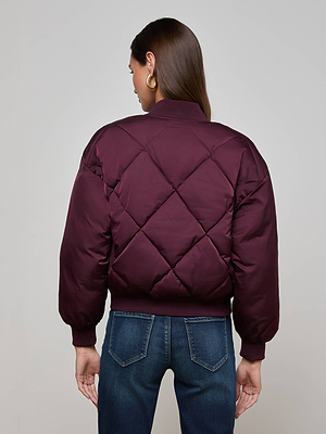A woman with long brown hair and gold hoop earrings models the L'AGENCE Violeta Reversible Puffer, shown in maroon, paired with blue jeans. She stands facing away from the camera against a plain gray background.