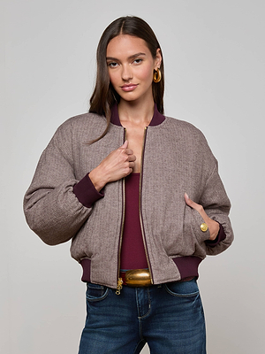 A woman with long brown hair wears the L'AGENCE Violeta Reversible Puffer in mauve over a maroon top and blue jeans. She stands facing forward, holding the jacket collar with one hand and has a neutral expression.
