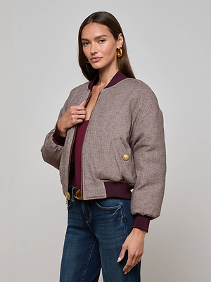 A woman with long brown hair wears the L'AGENCE Violeta Reversible Puffer, styled with a burgundy top, blue jeans, and gold hoop earrings, standing against a plain light gray background.