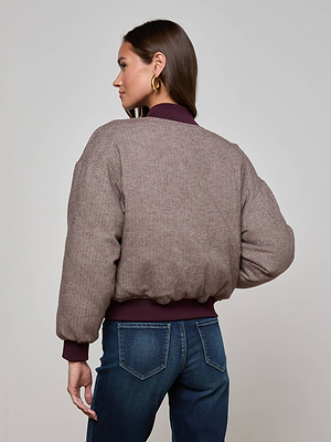 A woman with straight brown hair models the L'AGENCE Violeta Reversible Puffer, featuring quilted mauve satin with maroon cuffs and collar, gold hoop earrings, and blue jeans, as she faces away against a plain background.