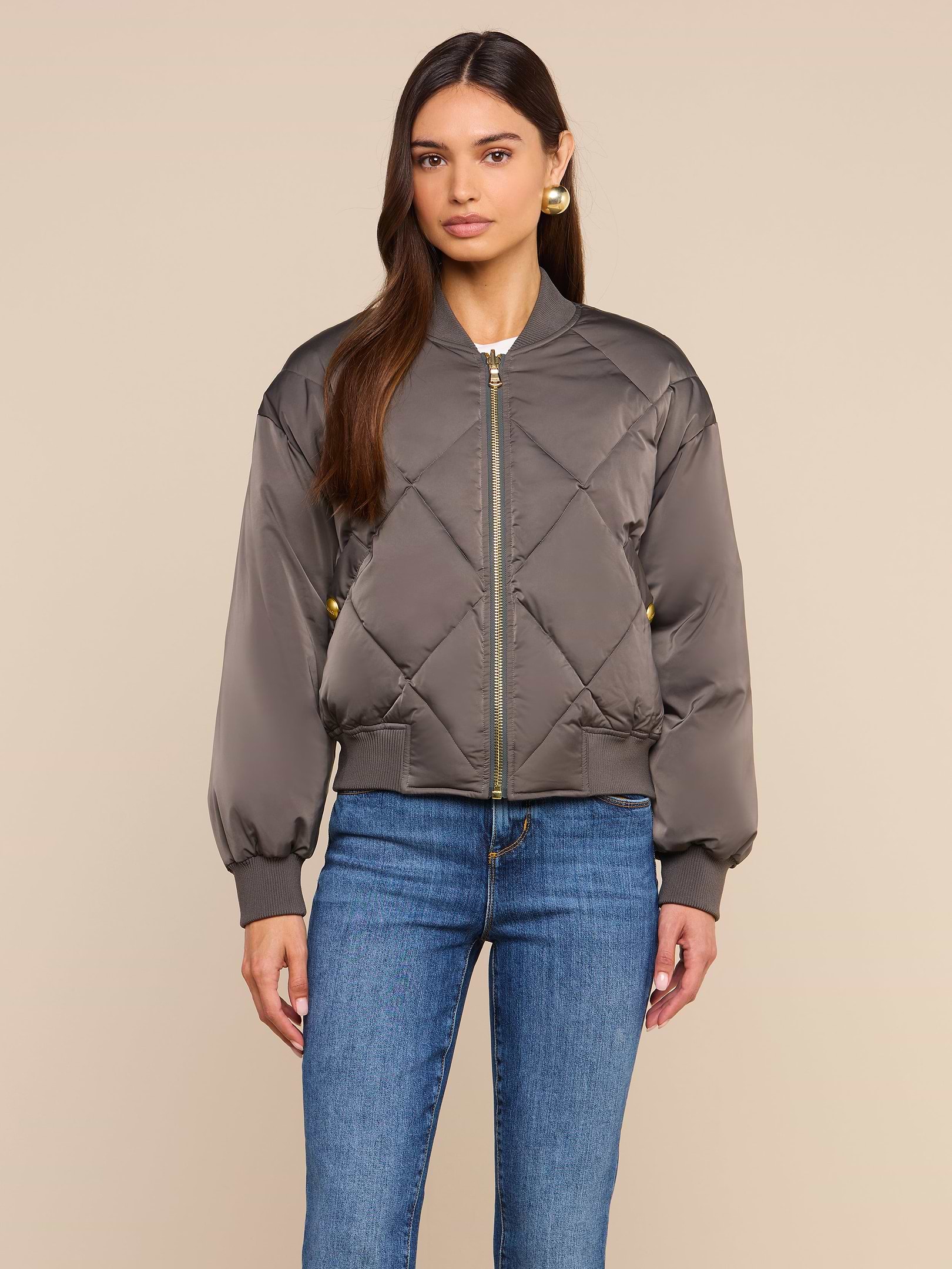 A woman stands against a plain background wearing the Violeta Reversible Puffer—a gray quilted satin bomber jacket with a front zipper—paired with blue jeans, looking at the camera with a neutral expression.