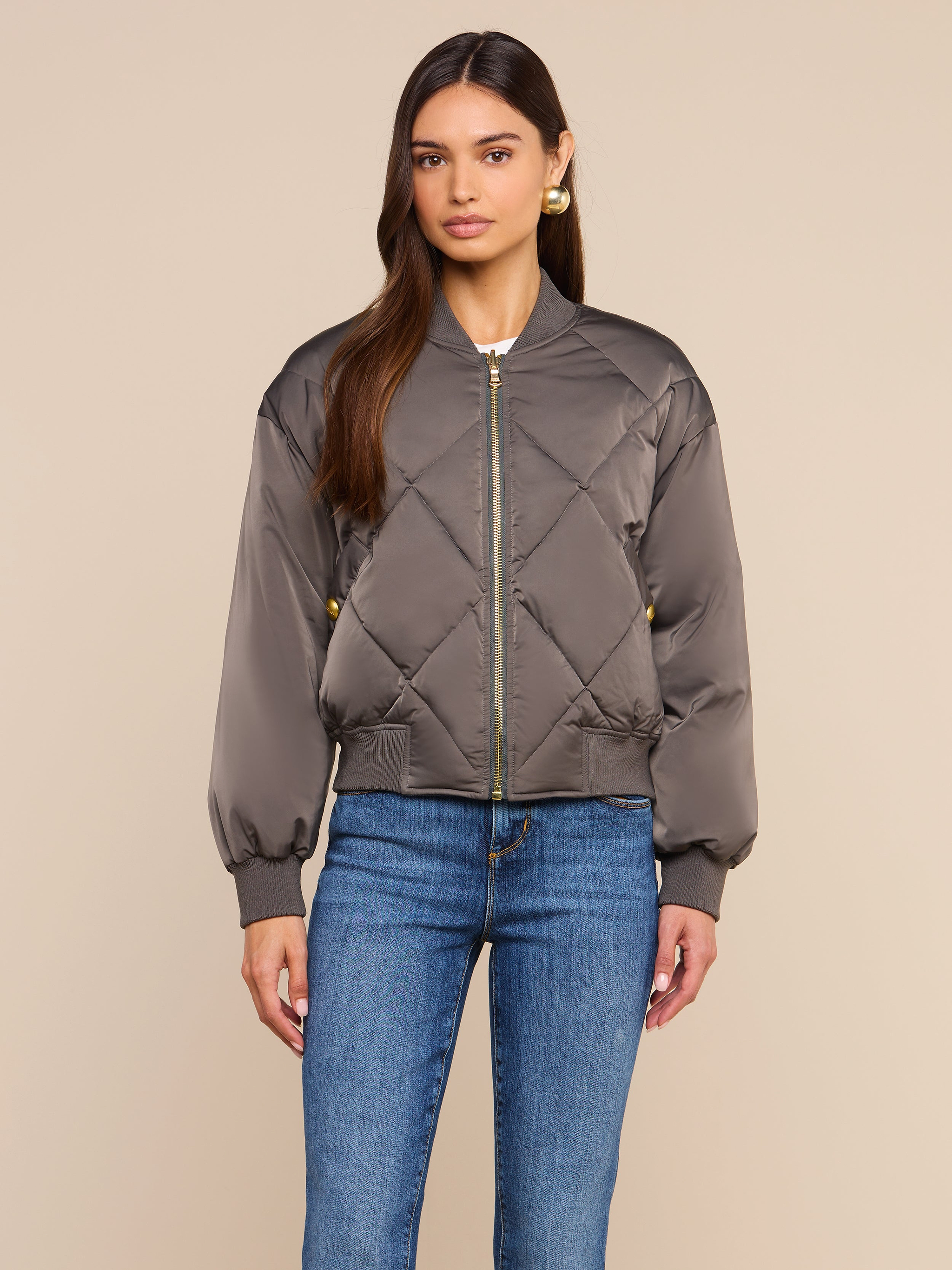 A woman stands against a plain beige background, facing forward with a neutral expression, wearing the Violeta Reversible Puffer in gray quilted satin and blue jeans.