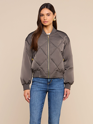 A woman stands against a plain beige background, facing forward with a neutral expression, wearing the Violeta Reversible Puffer in gray quilted satin and blue jeans.