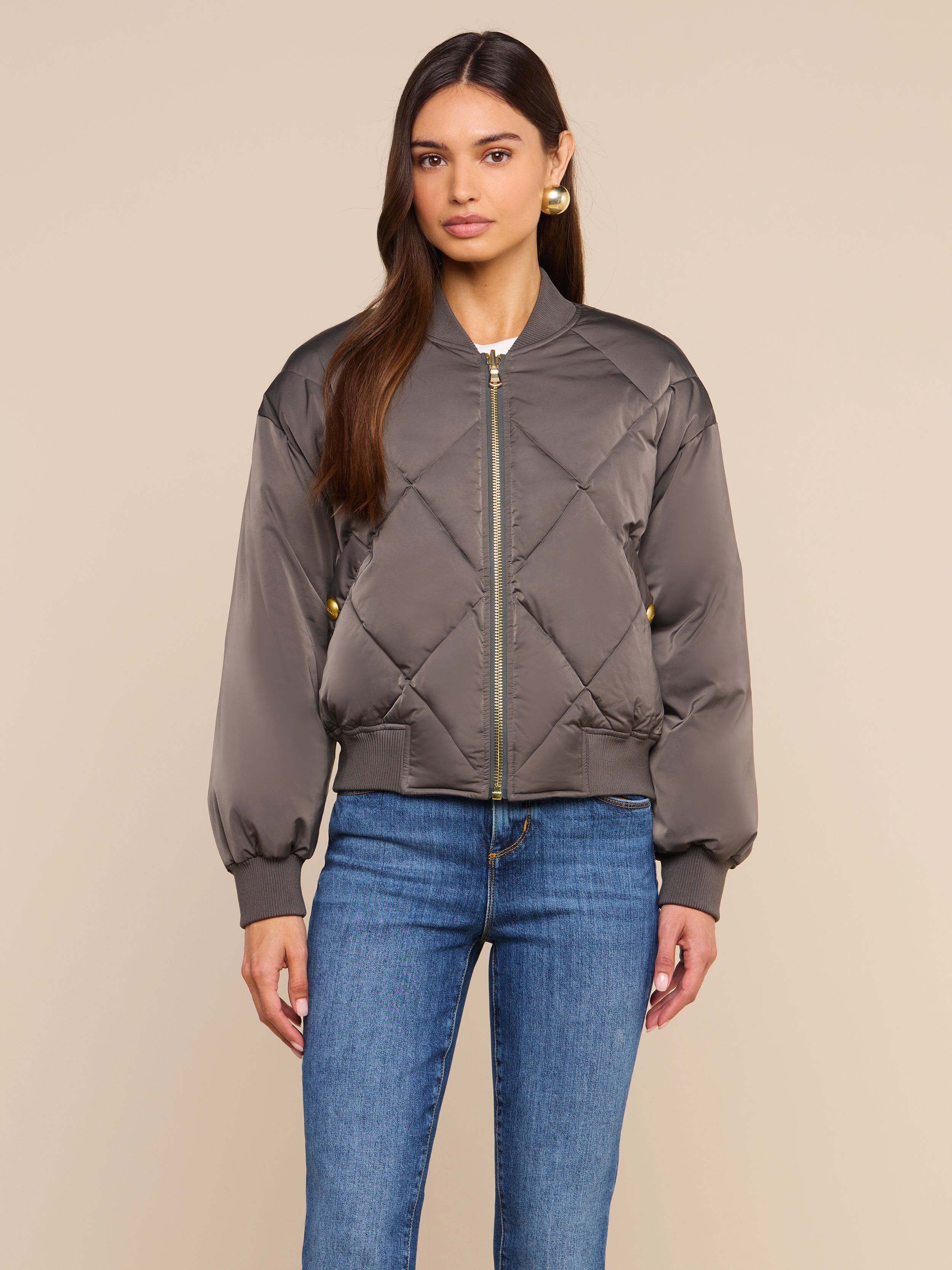 A woman stands against a plain beige background, facing forward with a neutral expression, wearing the Violeta Reversible Puffer in gray quilted satin and blue jeans.