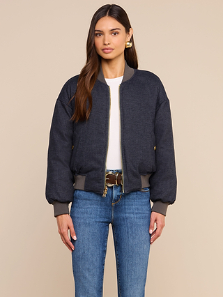 A woman with long brown hair wears the Violeta Reversible Puffer over a white top, paired with blue jeans and a brown belt. She stands against a plain beige background, facing forward with a neutral expression.