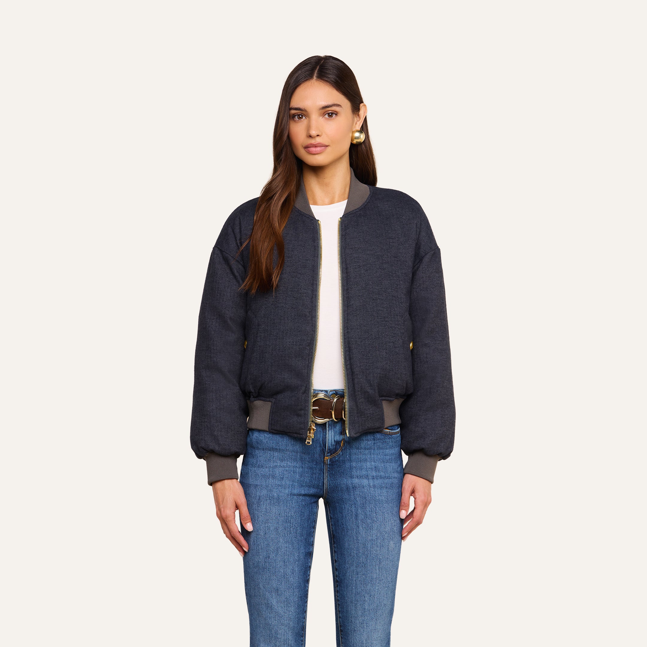 A woman with long brown hair wears the Violeta Reversible Puffer over a white top, paired with blue jeans and a brown belt. She stands against a plain beige background, facing forward with a neutral expression.