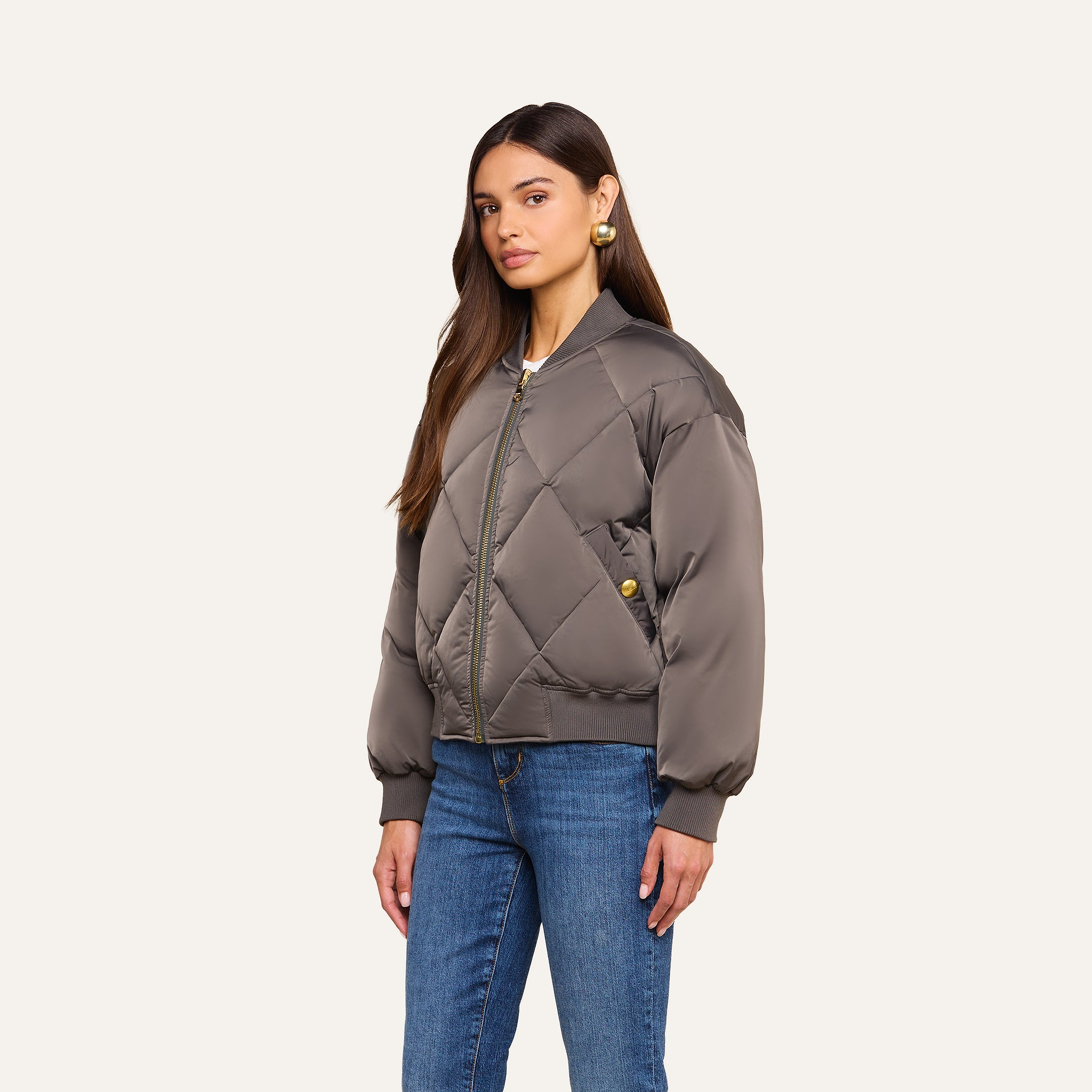 Woman with long brown hair wearing the Violeta Reversible Puffer in gray, styled with blue jeans and gold earrings, standing against a plain beige background.