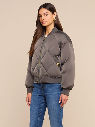 Woman with long brown hair wearing the Violeta Reversible Puffer in gray, styled with blue jeans and gold earrings, standing against a plain beige background.