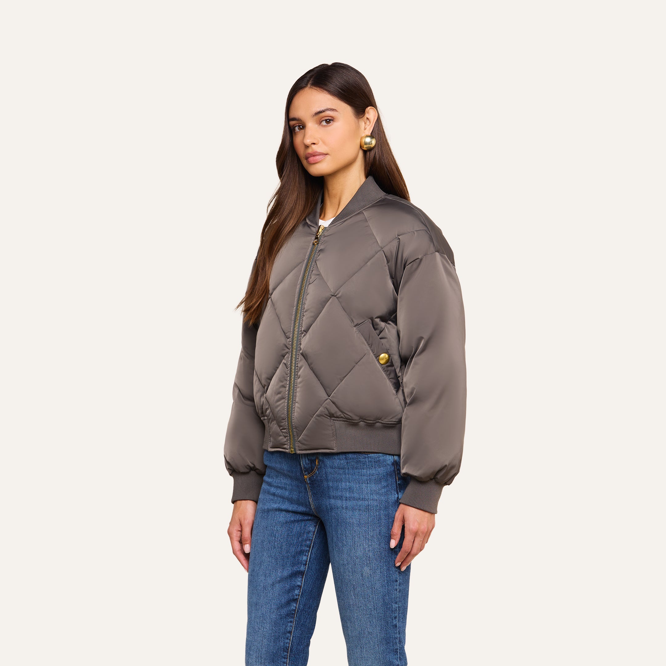 Woman with long brown hair wearing the Violeta Reversible Puffer in gray, styled with blue jeans and gold earrings, standing against a plain beige background.