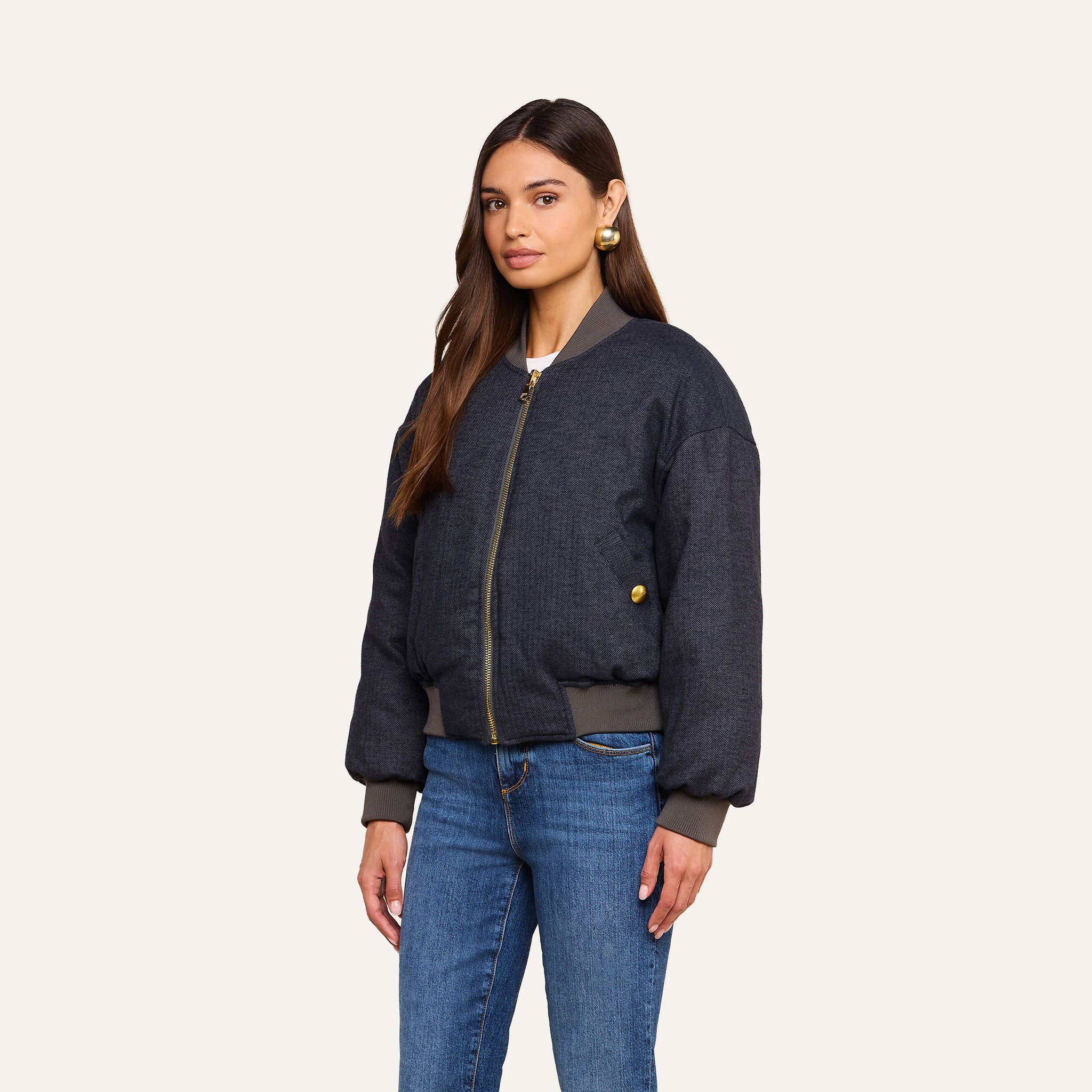 A woman with long brown hair wears the Violeta Reversible Puffer in dark gray, paired with blue jeans and gold earrings, standing against a beige background.