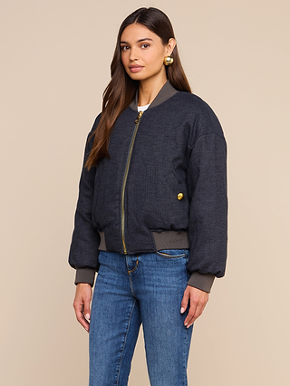 A woman with long brown hair wears the Violeta Reversible Puffer in dark gray, paired with blue jeans and gold earrings, standing against a beige background.