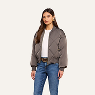 A woman wearing the Violeta Reversible Puffer, a gray quilted satin bomber jacket, over a white top and blue jeans stands against a beige background.