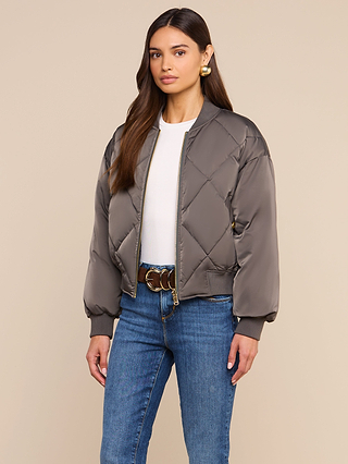 A woman wearing the Violeta Reversible Puffer, a gray quilted satin bomber jacket, over a white top and blue jeans stands against a beige background.
