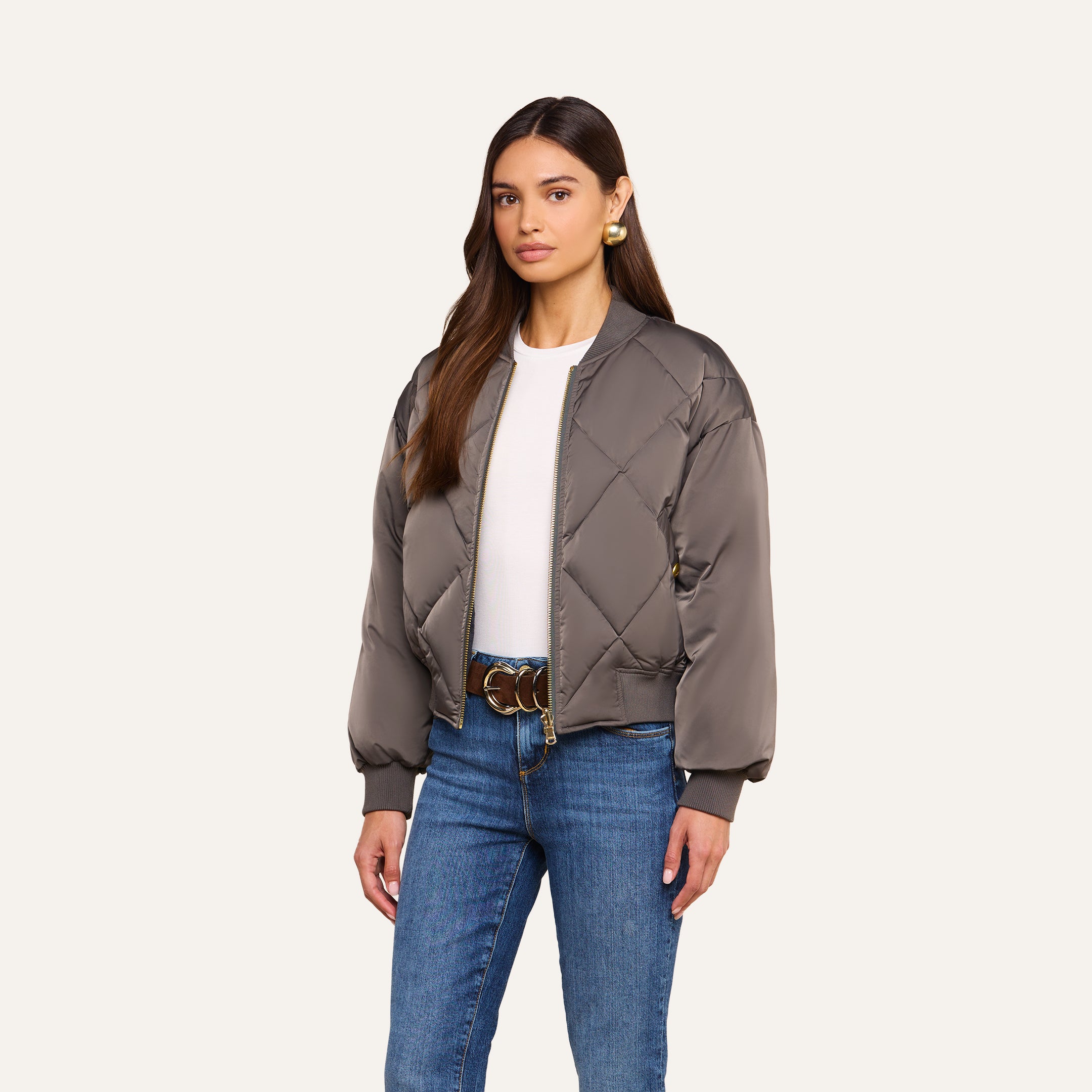 A woman wearing the Violeta Reversible Puffer, a gray quilted satin bomber jacket, over a white top and blue jeans stands against a beige background.